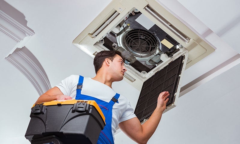 HVAC technician replacing a dirty air filter in a ceiling-mounted AC unit while holding a toolbox.