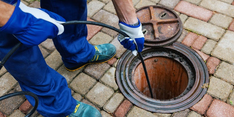 A person in blue work clothes and gloves using a sewer snake to clear a drain through an open manhole.