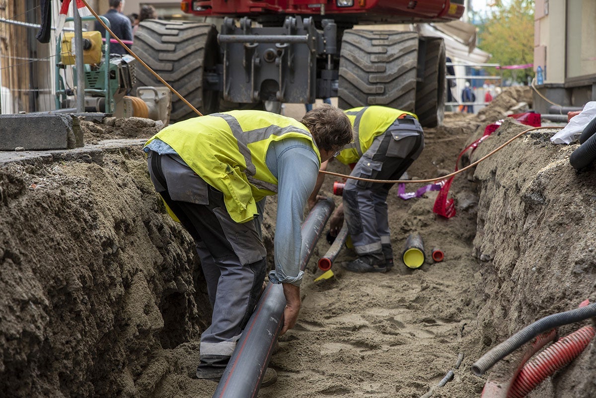 Two construction workers in high-vis vests laying pipes in a trench, with an excavator in the background.