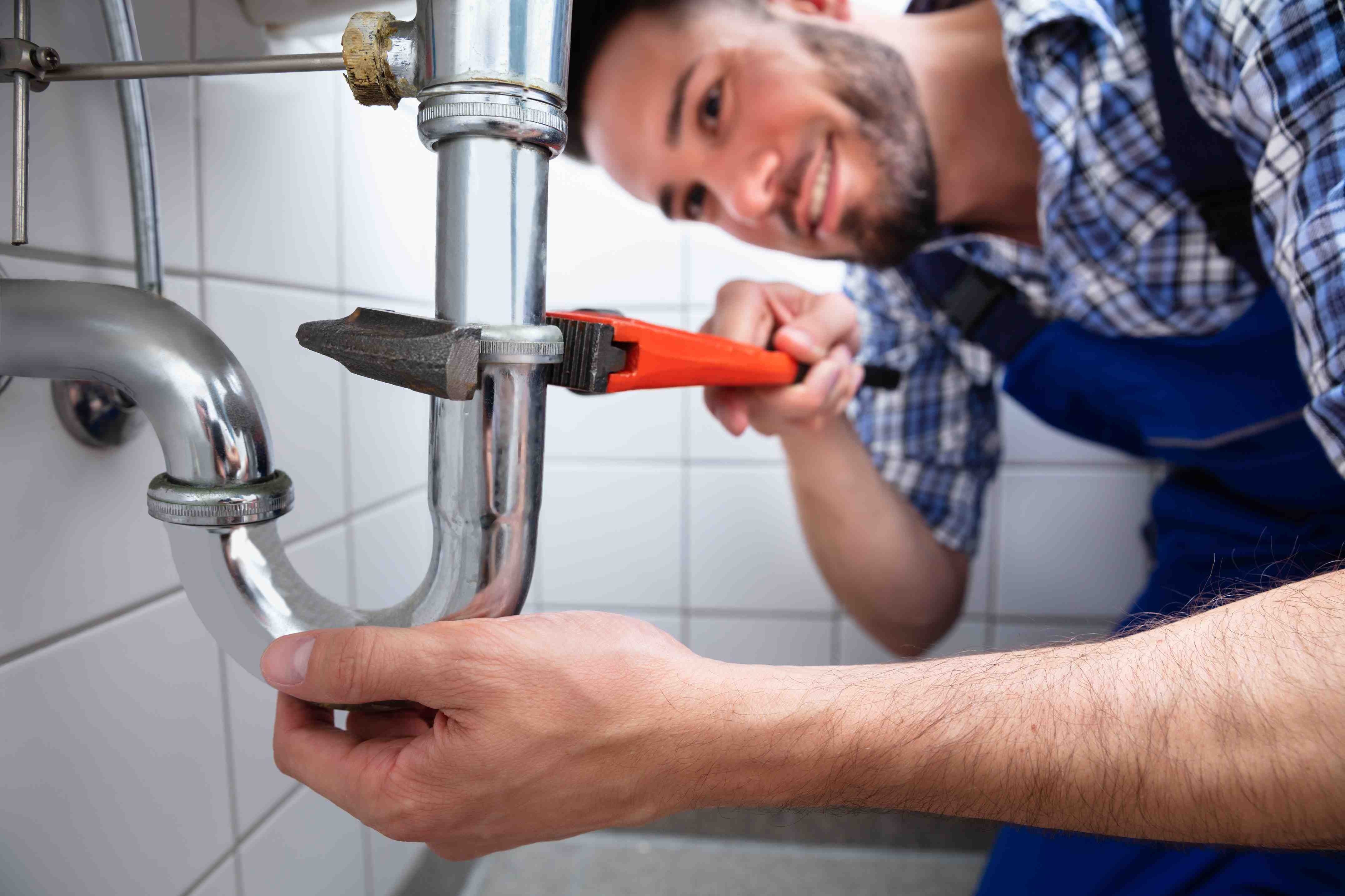 A smiling plumber in a blue plaid shirt and overalls tightens a pipe under a sink with a red wrench.