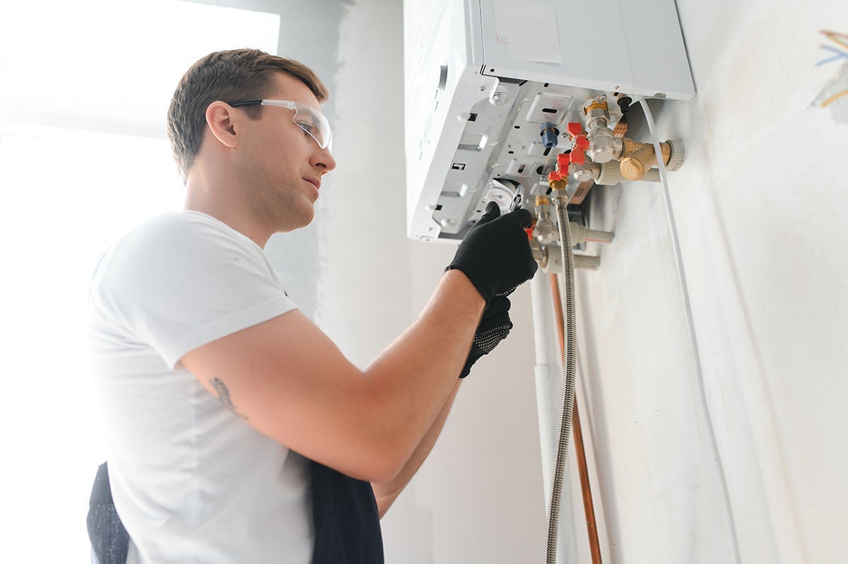 A man in safety glasses and work gloves, working on a gas boiler attached to a white wall.