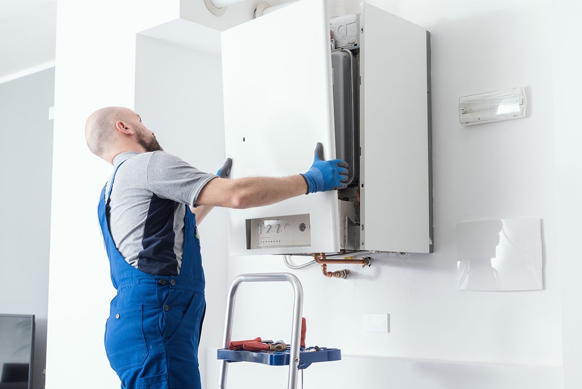 A technician in blue overalls and gloves installing a white wall-mounted boiler in a bright room.